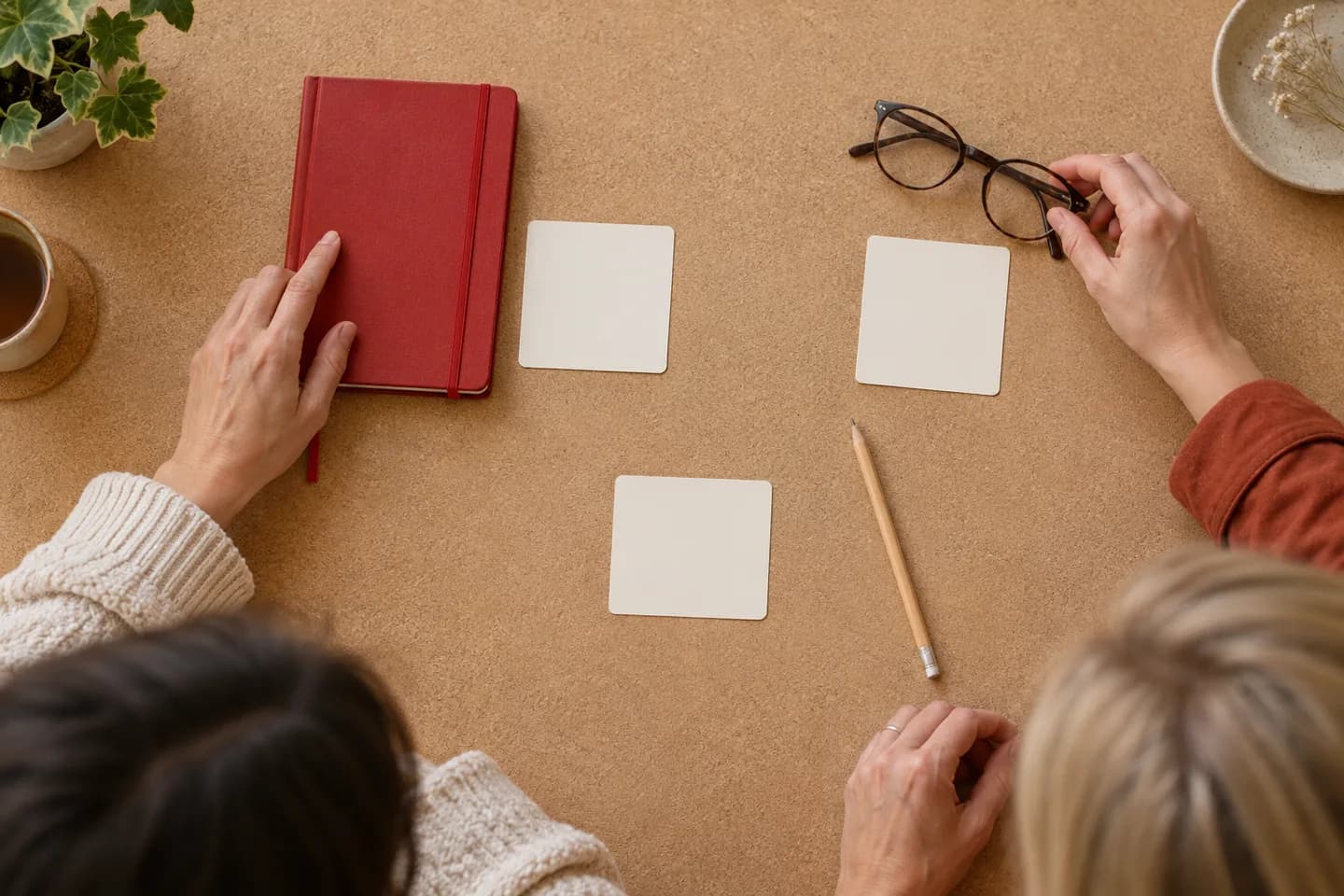 Deux mains placent un carnet rouge et des lunettes près de cartes vierges sur une surface en liège