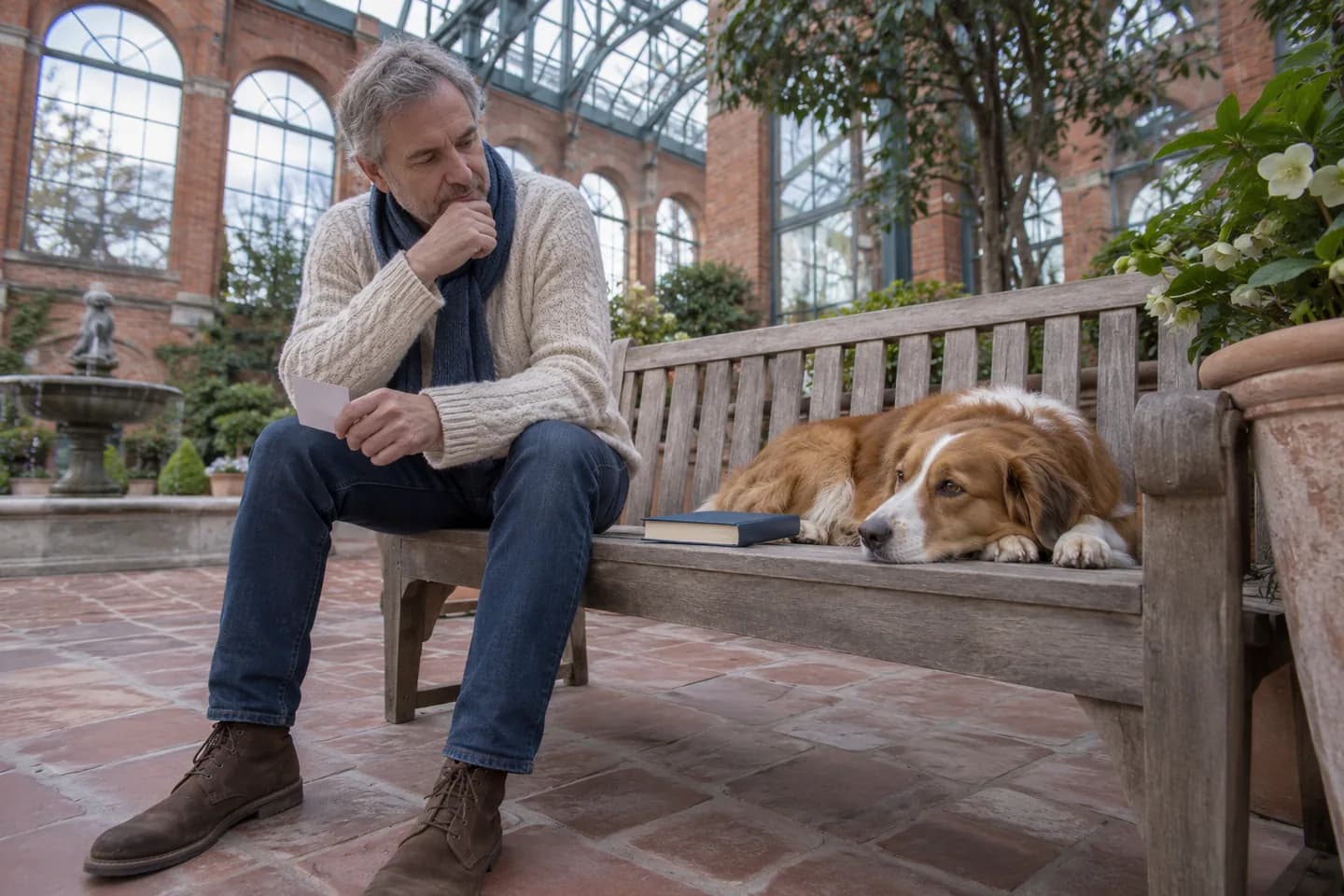 Hombre europeo observando un perro y un libro en un banco de un invernadero de ladrillo