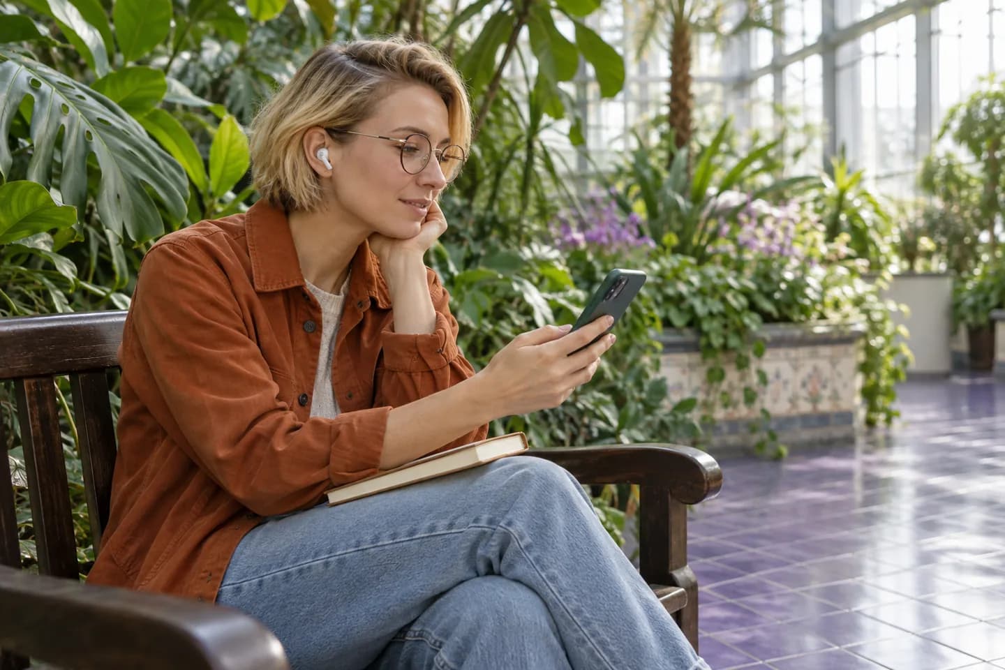European woman with earbuds listening on a conservatory bench beside tropical plants