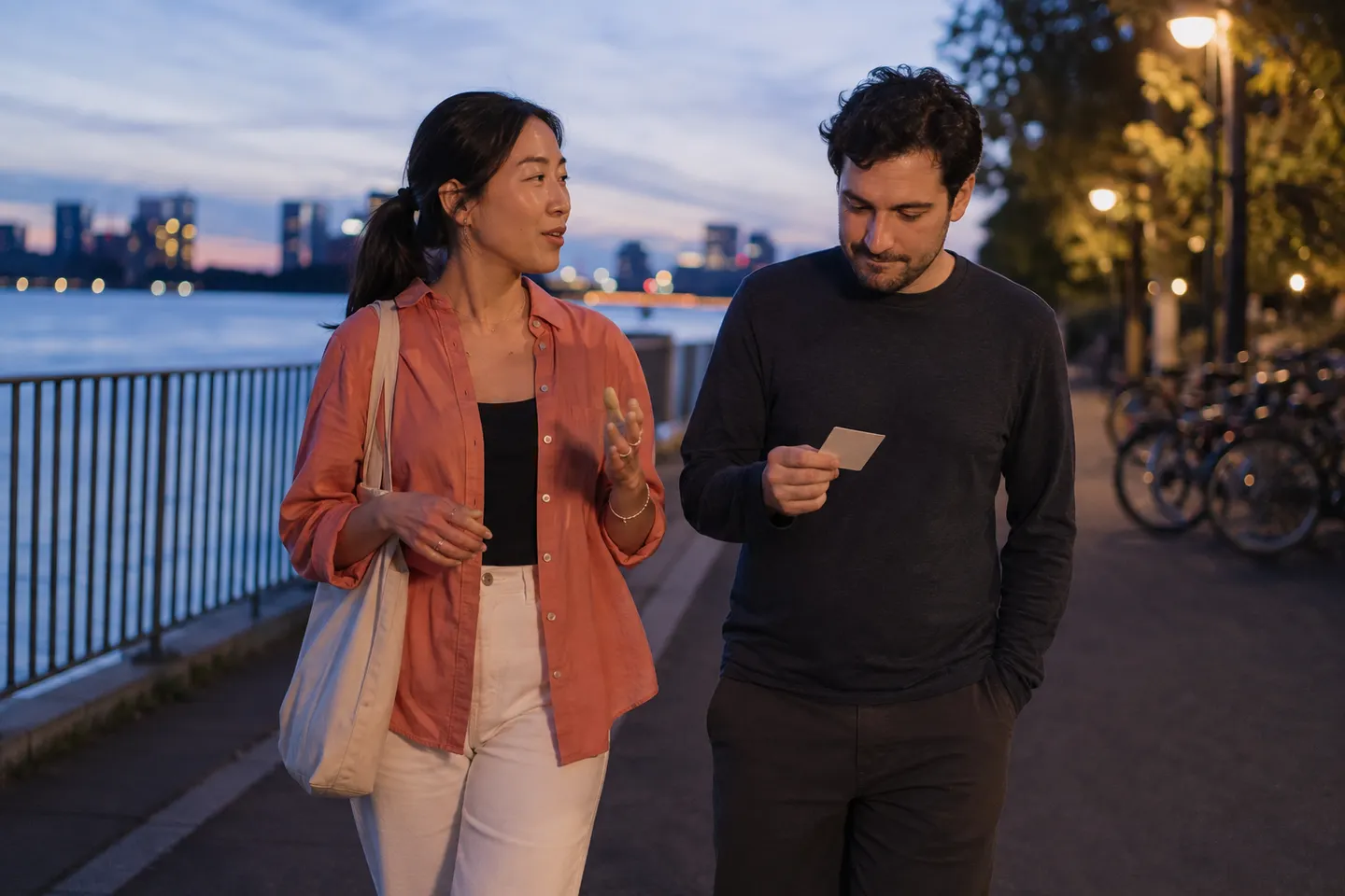 Two adult language partners walking beside a river while one answers a hidden question card from memory