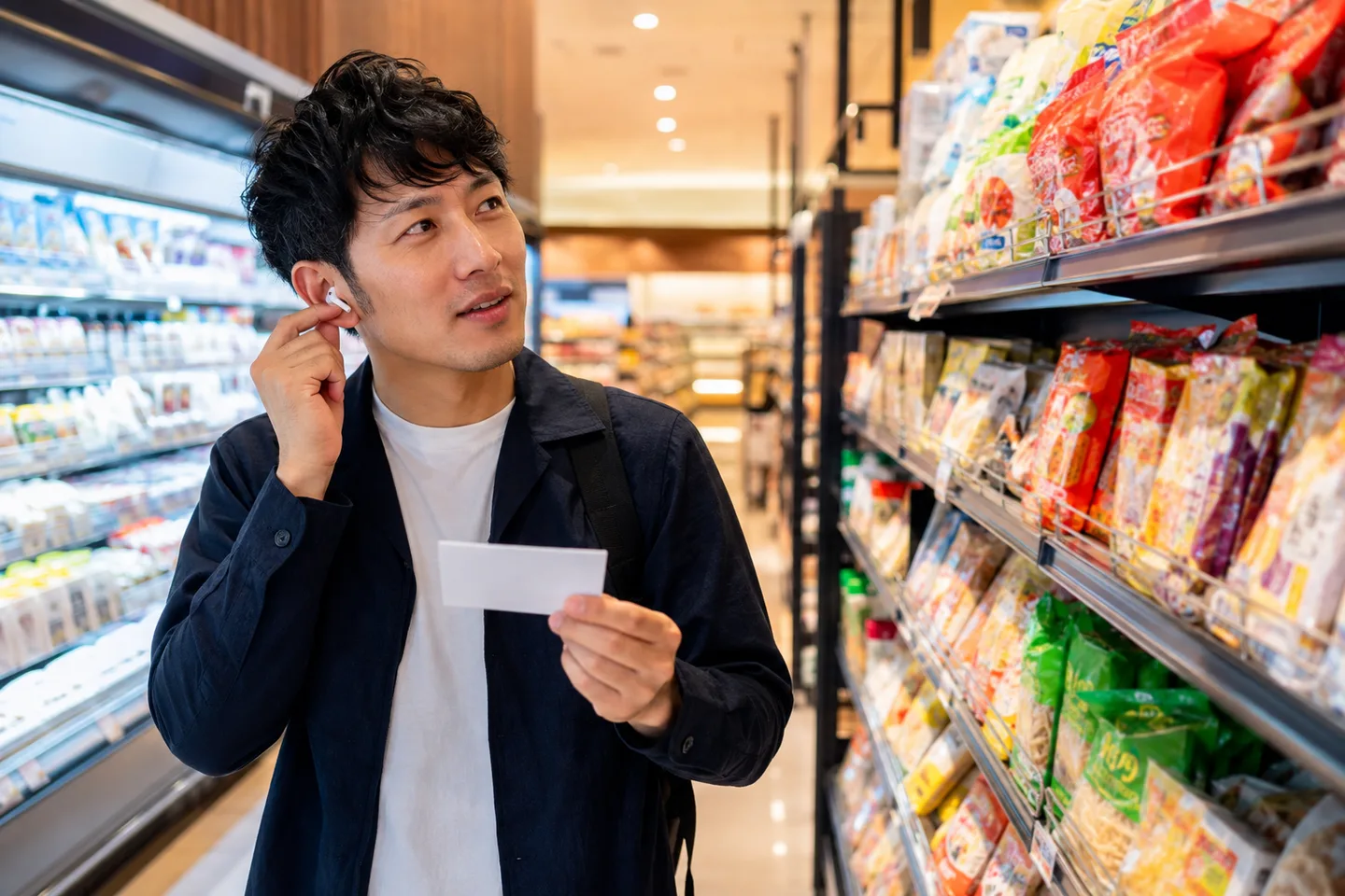 Estudiante adulto escucha préstamos escritos en katakana mientras compara productos cotidianos en un supermercado japonés luminoso